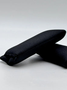 Zwei schwarze Schulterpolster für Sicherheitsgurte, in einer leicht schrägen Perspektive abgebildet, zeigen die Struktur des Materials.